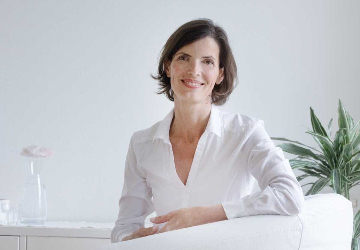 Portrait: Heilpraktikerin für Psychotherapie, Paartherapeutin und Kinesiologin Christina Pielken sitzt in ihrer Psychotherapie-Praxis in einem weißen Sessel, neben ihr steht auf der einen Seite eine Wasserkaraffe, auf der anderen eine Zimmerpflanze. Sie gibt Einzeltherapie und Paartherapie.